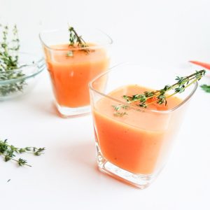 orange drink made with persimmon, carrot, thyme, lemon, honey; in a glass with thyme sprig on top on white surface
