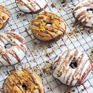 chocolate donuts iced with white chocolate and peanut butter, drizzled with milk chocolate, topped with almonds and pistachios on a wire rack