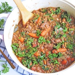 Kale, Lentil, and Beef Stew with carrots and parsley on top, in a pot with a wooden spoon