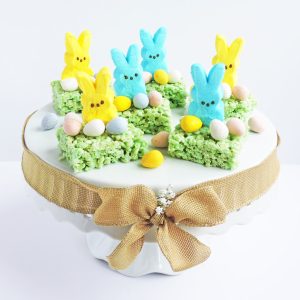 green rice cereal treats topped with yellow and blue marshmallow bunnies and candy eggs, sitting on a white pedastal for Easter