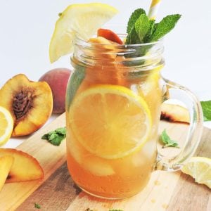 Bourbon Ginger Sweet Peach Tea in a mason jar with mint leaves, ice, lemon slices, and sliced peaches, with a striped yellow straw on a wooden board