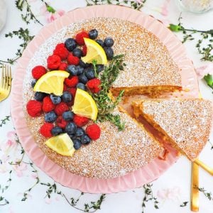 Rustic Lemon & Thyme Olive Oil Cake on a pink pedestal cake stand topped with raspberries, blueberries, lemon wedges, thyme, and powdered sugar, on a white surface with Spring pink flowers