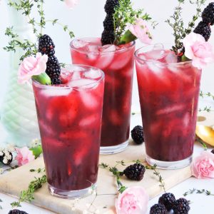 Peachy Blackberry & Thyme Cocktail served in a glass of ice topped with a pink flower, blackberry skewer, and fresh thyme. perfect for Spring, a bridal shower, wedding or mother's day