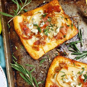 Mini Pear, Prosciutto, & Gorgonzola Flatbread Pizzas topped with rosemary and balsamic vinaigrette on a baking sheet